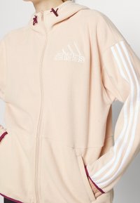 Světle růžový fleecový zip-up hoodies s kapucí na šňůrku, bílým vyšitým logem a kontrastními bílými pruhy po rukávech.