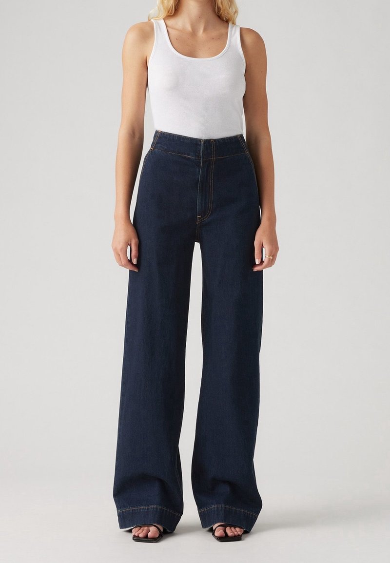 Femme portant un top blanc sans manches, un jean bleu foncé taille haute à jambes larges et des sandales noires à bout ouvert, debout devant un fond uni.