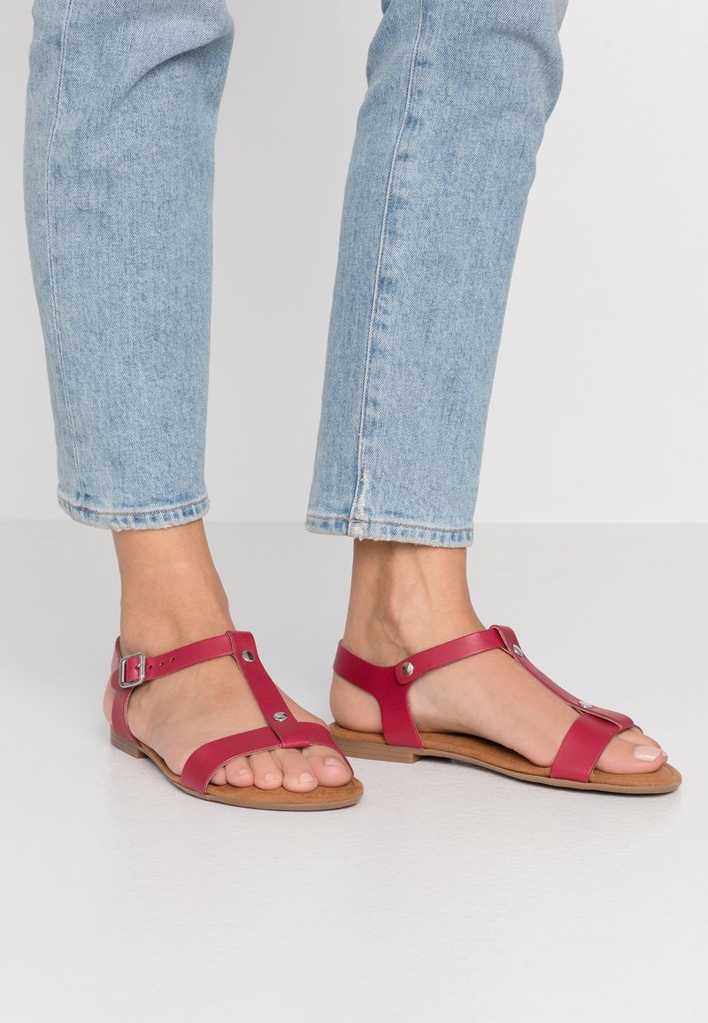 Pieds chaussés de jeans bleu clair et de sandales plates rouges avec brides autour de la cheville et clous métalliques, debout sur un sol gris clair.
