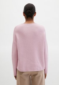 Femme aux cheveux foncés attachés en chignon, portant un pull ample en maille violet clair et un pantalon beige, vue de dos sur un fond blanc uni.