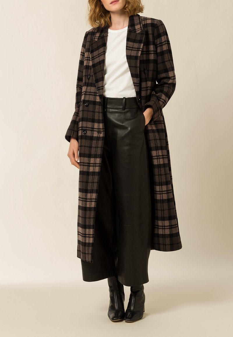 Femme portant un long manteau marron et noir à carreaux, un haut blanc, un pantalon large en cuir noir et des bottes à talons noires, debout avec une main dans la poche.