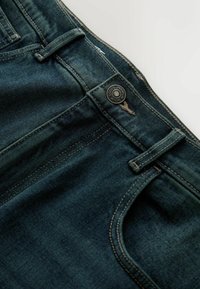 Tumši zila džinsa bikses ar gludu tekstūru, vieglu izbalēšanu, metāla pogas aizdari un dzeltenām šuvēm gar kabatām un šuvēm.