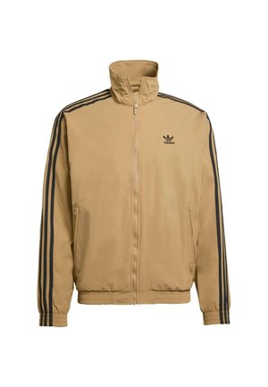Lichtbruine ritsjack gemaakt van stof met een gladde textuur. Bevat zwarte strepen op de schouders en mouwen, met een geborduurd logo.