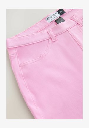 Przycięte różowe legginsy z dżerseju o gładkiej strukturze, z przeszytym paskiem w talii i przednimi kieszeniami; etykieta zawiera informacje o rozmiarze i pielęgnacji.