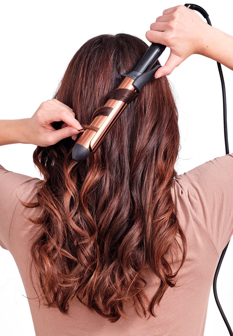 Prostownica do włosów z różowo-złotą rurką, czarną rączką i gumowym końcówką, kręcąca ciemno-brązowe, falowane włosy, z rękami umieszczonymi na urządzeniu.
