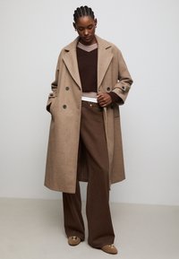 Beige double-breasted wool coat with wide lapels, front pockets, and black buttons, layered over a brown sweater and brown wide-leg pants.