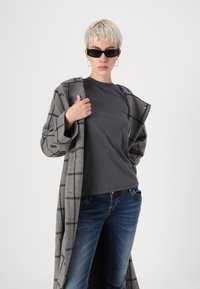 Person with short blonde hair wearing black sunglasses, hoop earrings, gray checkered coat, dark gray t-shirt, and blue jeans, posing confidently.