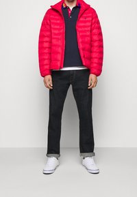 Röd quiltad pufferjacka, marinblå polo t-shirt, mörka jeans och vita sneakers. Jackan har hög krage, blixtlås och ribbade ärmslut.