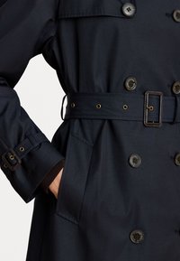 Tmavě modrý trench coat s páskem v pase, dvojitými knoflíky, bočními kapsami a nastavitelnými manžetami. Vyrobeno z hladkého materiálu s minimální texturou.