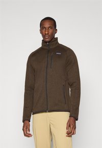 Brown fleece jacket with a high collar and full zip, featuring side pockets and a small logo on the chest. Smooth texture, fitted design.