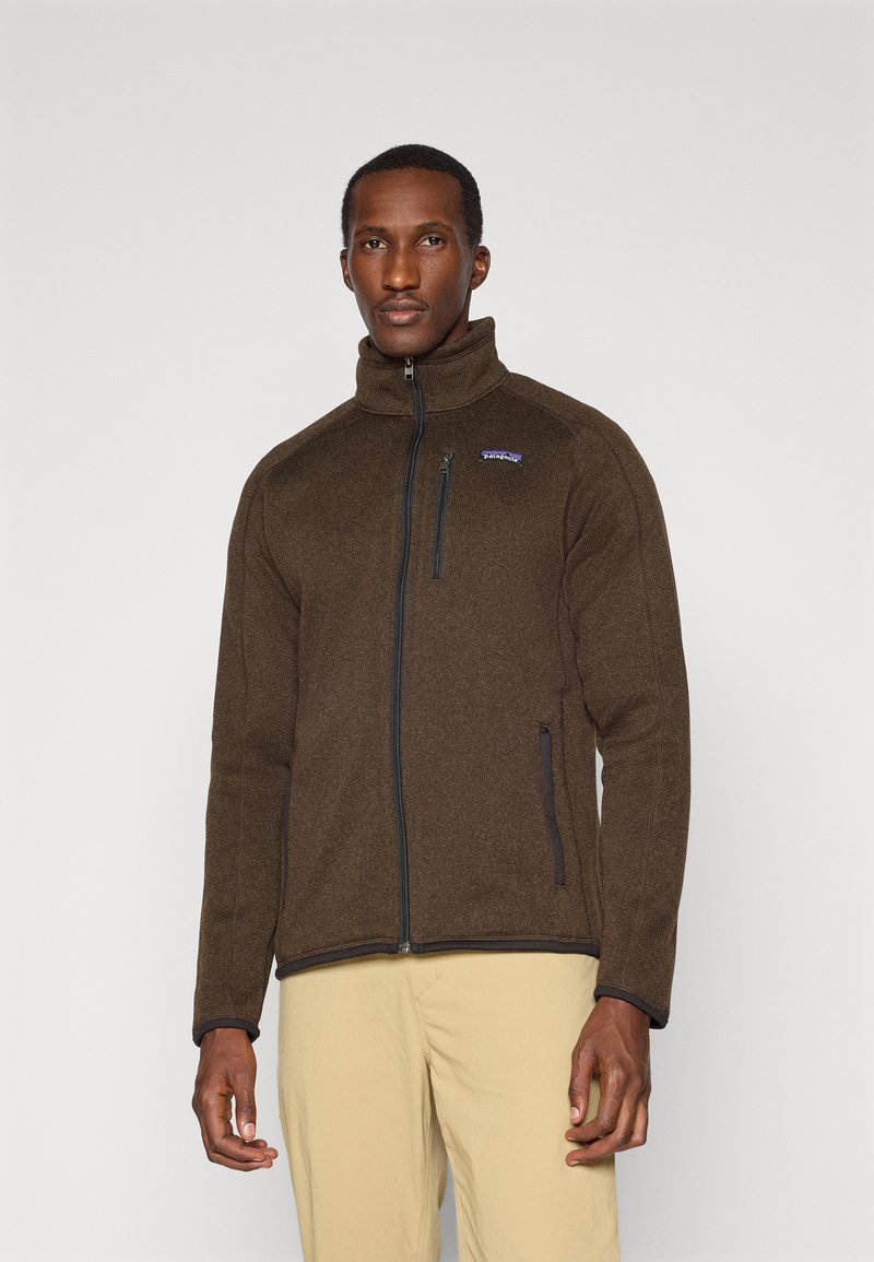Brown fleece jacket with a high collar and full zip, featuring side pockets and a small logo on the chest. Smooth texture, fitted design.