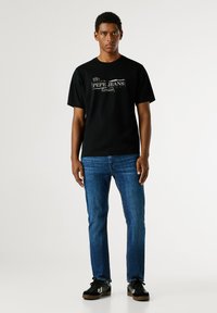 Černé bavlněné tričko s bílou potiskem loga, kombinované s modrými džíny. Model má na sobě černé tenisky s bílými akcenty.