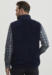 Tamno plavi flis prsluk s visokim ovratnikom, mekan na dodir i bez vidljivih zatvarača. Nositi preko kariranog plavog i sivog dugog rukava.