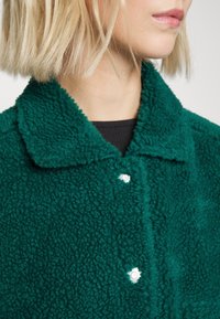 Grön, fluffig fleecejacka med krage, prydd med silverfärgade tryckknappar och en texturerad yta. Närbild av tyget och knapparna.