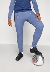 Blå Nike joggebukser med et Paris-logo, tætsiddende design, med manchetter ved anklerne, parret med sorte sportssko på en mønstret fodbold.