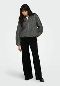 Veste grise texturée avec fermeture éclair à l'avant, taille élastique et manches bouffantes, associée à un pantalon noir à jambes larges et des chaussures plates.