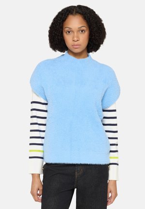 Woman with curly hair wearing a light blue fuzzy short-sleeve sweater over a long-sleeve white shirt with navy blue stripes and black pants.