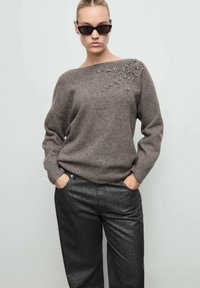 Suéter gris con escote barco y detalles de joyas en un hombro, combinado con unos pantalones negros brillantes.