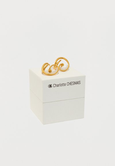 Ζευγάρι χρυσών σπειροειδών σκουλαρικιών τοποθετημένων επάνω σε ένα λευκό κουτί κοσμημάτων της Charlotte Chesnais, με φόντο απλό.