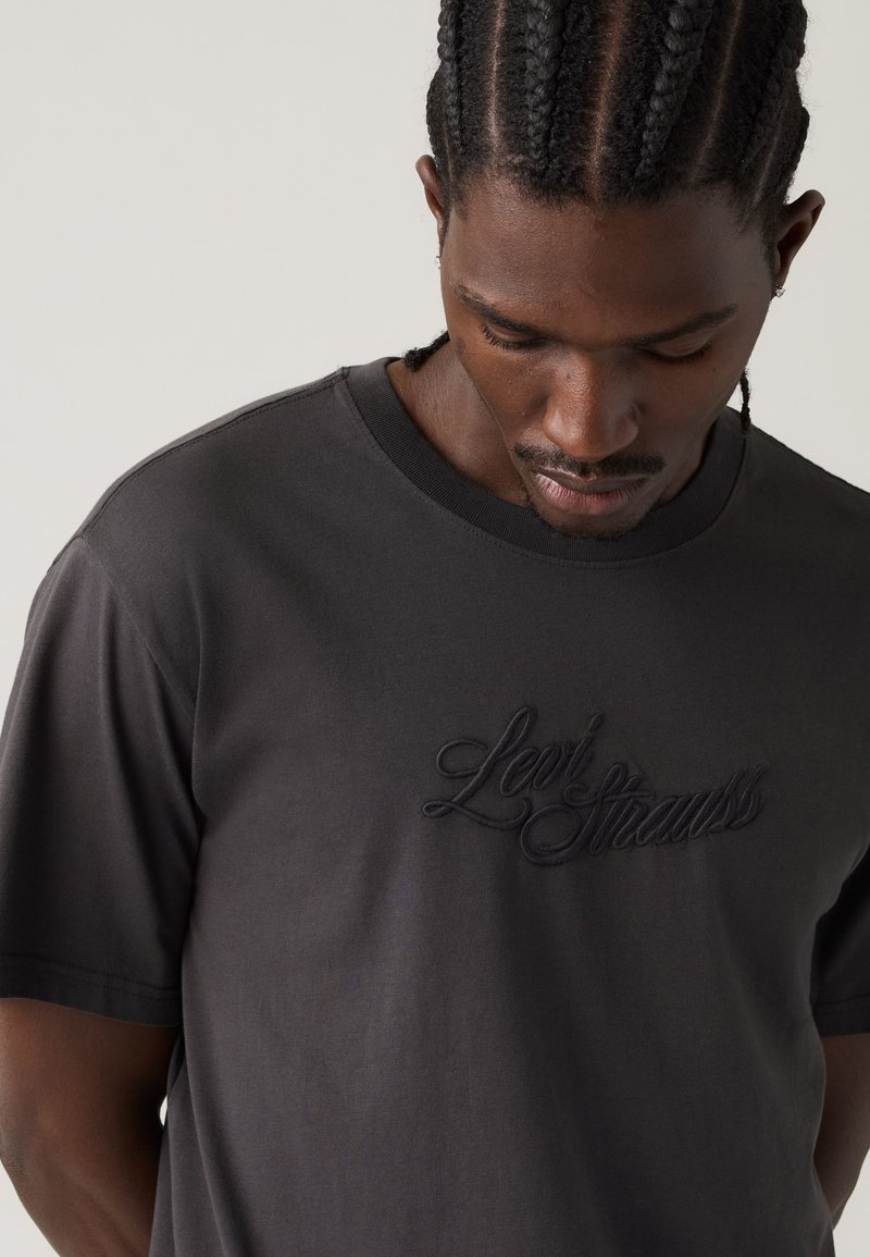 Man with braided hair wearing a dark Levi Strauss t-shirt looking down against a plain light background.