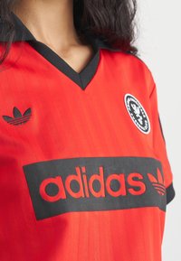 Czerwona koszulka piłkarska z czarnym kołnierzykiem, z pionowymi paskami, czarnym logo "adidas" na przodzie oraz okrągłą naszywką na klatce piersiowej.