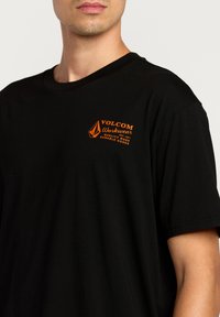 Sort bomulds t-shirt med et orange trykt logo, der står "VOLCOM Workwear" på venstre bryst. Enkel rund halsudskæring, korte ærmer.