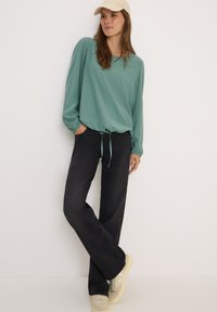 Blusa verde chiaro a maniche lunghe con strisce verticali, aderente in vita con un fiocco. Abbinata a pantaloni neri a gamba larga e sneakers beige.