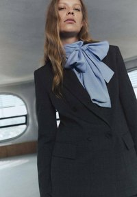 Ciemnoszary blazer w kratkę z podwójnymi guzikami, w połączeniu z jasnoniebieską dużą muszką. Gładka tkanina i wyraźna forma.