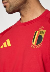 Rdeča športna majica z zlatim in črnim grbom belgijske reprezentance na prsih ter rumenim logotipom Adidas na levem rami.