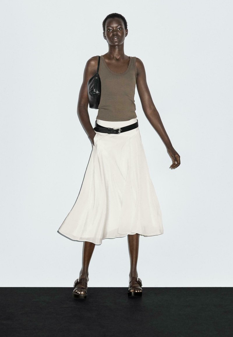 Bruine tanktop, witte uitlopende rok, zwarte riem en zwarte schoudertas. Donkerbruine sandalen met decoratieve hardwaredetails. Effen achtergrond.