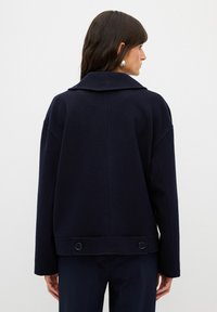 Jacket z vlněné směsi v námořnické modři s širokým límcem, spadlými rameny a knoflíkovými detaily na lemu. Jednoduchá, kvadratická silueta s texturovanou tkaninou.