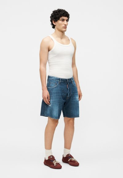 Νέος άνδρας που στέκεται φορώντας ένα λευκό φανελάκι, μπλε τζιν σορτς, μπεζ κάλτσες και κόκκινα αθλητικά παπούτσια μπροστά σε απλό λευκό φόντο.