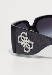 Gafas de sol negras con lentes degradados, que presentan un costado texturizado adornado con un logo de strass plateado en un diseño entrelazado.