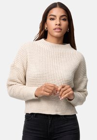 Beżowy sweter z dzianiny z wysokim, prążkowanym kołnierzem. Posiada strukturalny wzór, krótszy krój i prążkowane mankiety. Noszony z czarnymi dżinsami.