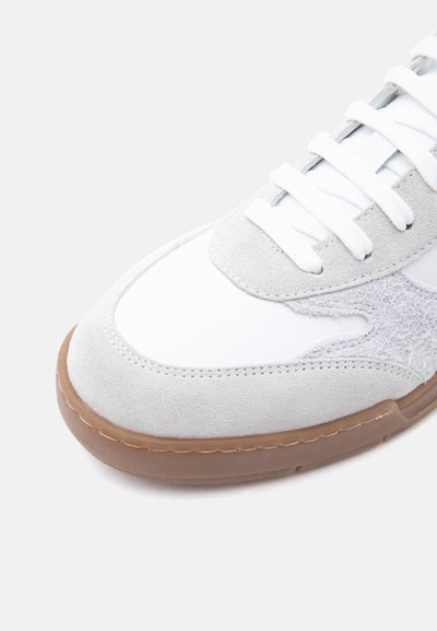 Λευκό αθλητικό παπούτσι με μπεζ σουέντ επικαλύψεις, λευκά κορδόνια, υφασμένη λεπτομέρεια στο πλάι και καφέ λαστιχένια σόλα σε απλό φόντο.