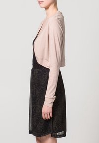 Ljusrosa kort kardigan med långa ärmar, kombinerad med en svart kjol med guldprickar. Slät textur och avslappnad passform.