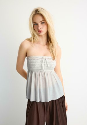 Plavuša u bijelom korzetu od čipke i nabranom topu, uparenom s širokim smeđim hlačama, stoji ispred jednobojne bijele pozadine.