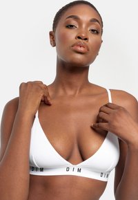 Bralette triangular blanco con tirantes finos y una banda elástica ancha que presenta la marca "DIM" en negro. Textura suave y diseño minimalista.