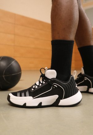 Jambes portant des chaussettes noires et des chaussures de basketball noires et blanches, debout sur un terrain couvert près d'un ballon de basketball noir.