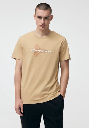 Młody mężczyzna z krótkimi włosami, ubrany w beżową koszulkę z napisem "różnorodność" i czarne spodnie, stoi na tle jednolitego, jasnego tła.