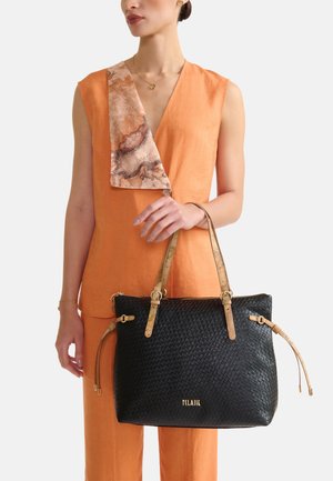 Donna con outfit arancione senza maniche che tiene una borsa tote intrecciata nera con manici color cuoio e logo dorato, indossando una sciarpa con motivo marmo sulla spalla.