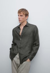 Donkergroen linnen button-up shirt met een ontspannen pasvorm en lange mouwen. Heeft een ondiepe halslijn en kleine knopen. Draagt met lichtgrijze jeans.