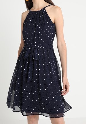 Robe bleu marine sans manches à pois blancs, avec un col licou et une ceinture nouée à la taille, portée par une femme debout.