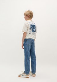 Hvid grafisk t-shirt med et blåt trykt design på ryggen, sammen med lyseblå denim jeans og cremefarvede sneakers med blå detaljer.