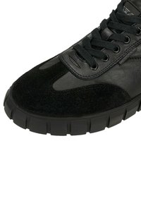 Zwarte sneaker met dikke geribbelde zool, suède neus, leren bovenwerk, zwarte veters en een klein merkteken "Wojas" aan de zijkant.