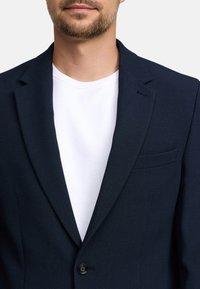 Blazer blu navy con finitura testurizzata, chiusura a un bottone e rever stretto; indossato sopra una semplice t-shirt bianca.