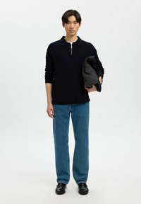 Maglione zip nero, sottoshirt grigio chiaro, jeans dritti blu e scarpe nere. Modello che tiene un oggetto grigio scuro. Sfondo semplice.