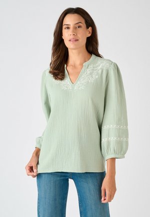 Donna che indossa una blusa verde chiaro con trama e ricamo floreale bianco sul collo e sulle maniche, abbinata a jeans blu, in piedi su sfondo bianco.