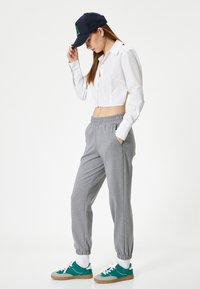 Camisa blanca recortada de botones, pantalones de chándal gris con cintura elástica y zapatillas verdes con detalles blancos. Lleva una gorra azul marino y pendientes de aro.