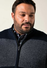 Mörkblå stickad ziptröja med en texturerad design och ljusgrå horisontella ränder. Bärs över en rutigt skjorta.